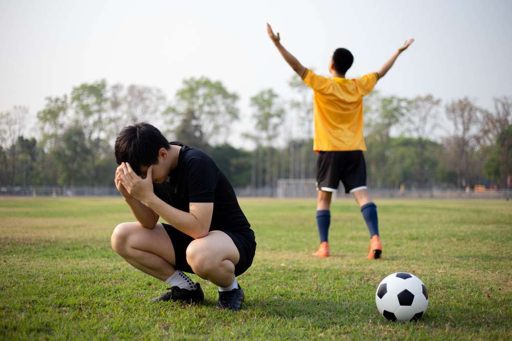 黒いユニフォームを着たサッカー選手が頭を抱えてしゃがみ込み、黄色いユニフォームを着た別の選手が腕を上げて勝利を祝っている。芝生に置かれたサッカーボールが、スペインでの試合の緊迫した感情を映し出している。