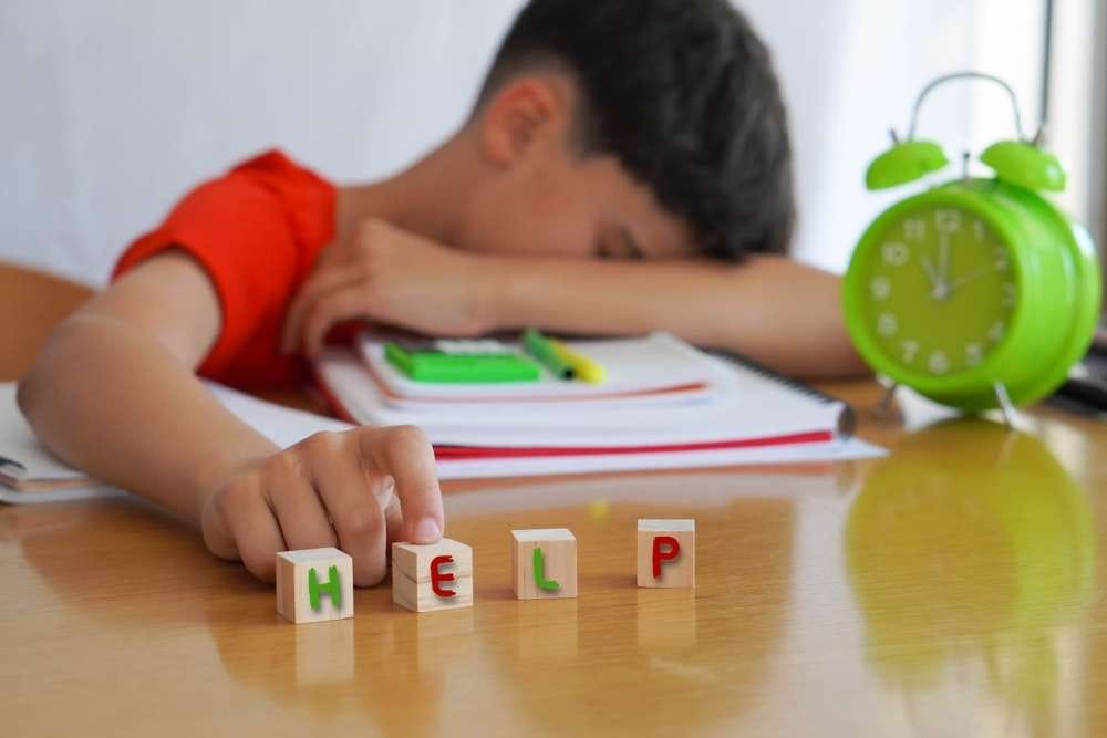 子どもが机に頭を乗せており、机の前にはブロックで「HELP」と書いてある。近くにはノート、緑色の目覚まし時計、壁にピンで留められたスペインの地図がある。