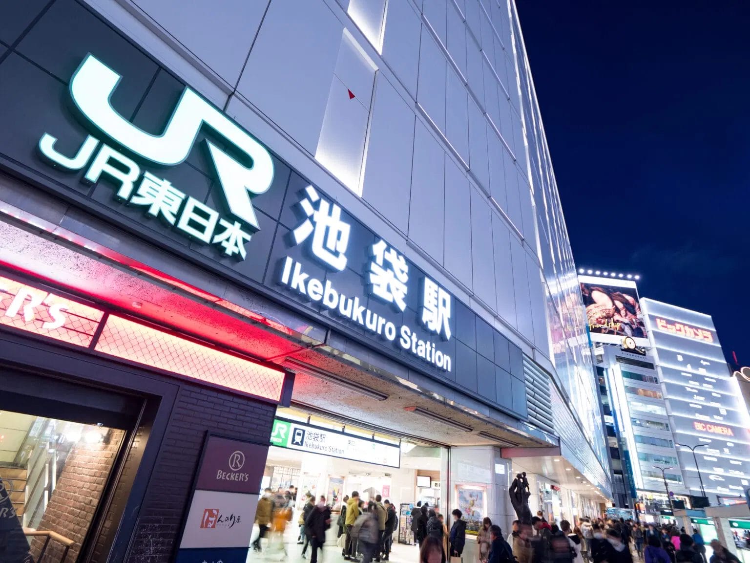 ライトアップされた東京・池袋駅の入り口を通り過ぎる群衆。モダンなビルのファサードには、日本語と英語のわかりやすいサインが掲げられている。.